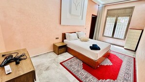 A bedroom with a bed, a nightstand, a wooden table, a red patterned rug, and a window with curtains.