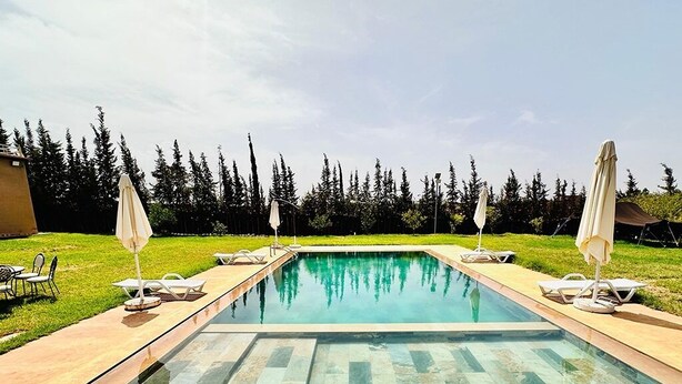 A rectangular swimming pool with a clear blue water surface, surrounded by a tiled deck. There are lounge chairs and umbrellas on both sides of the pool.