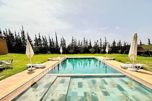 Et rektangulært swimmingpool med en klar, blå vandoverflade, omgivet af en flisebelagt terrasse. Der er liggestole og parasoller på begge sider af poolen.