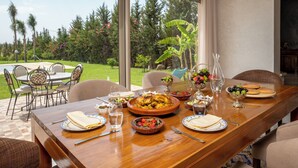 A dining table set with plates, cutlery, and various dishes, overlooking a garden with chairs and a table outside.