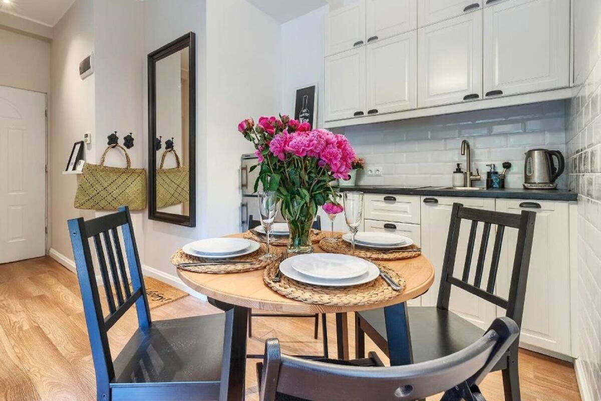A compact kitchen with white cabinets, a round wooden table set for four, and a vase of pink flowers.