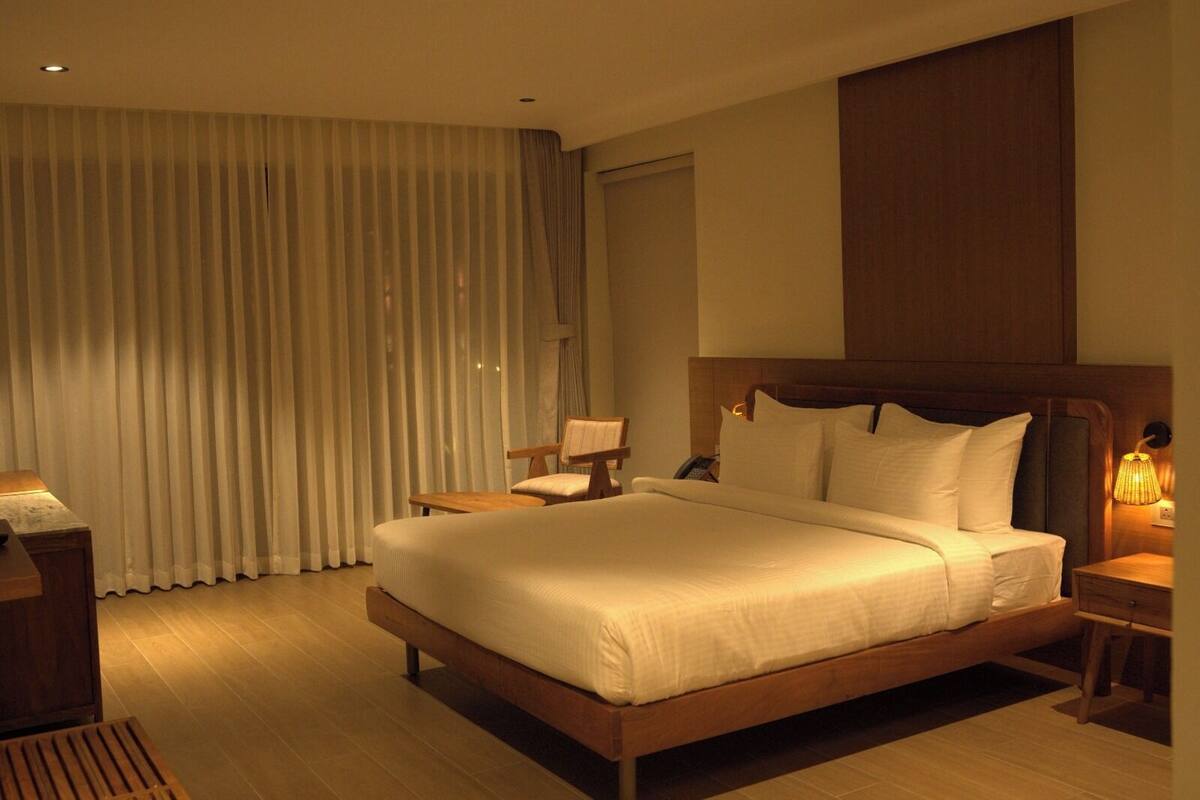 Habitación de hotel con una cama grande, cabecero de madera, un escritorio con silla, una mesita de noche con lámpara y una ventana con cortinas.