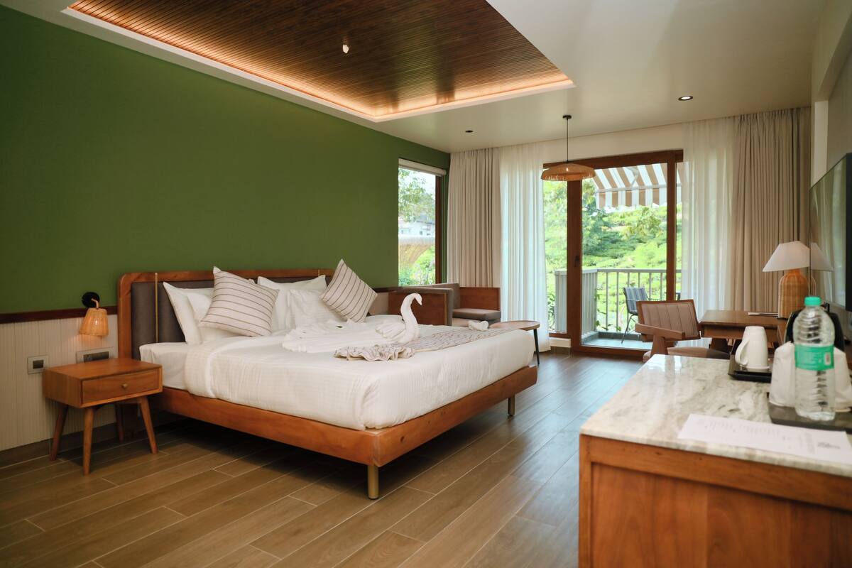Habitación de hotel con una cama grande, una mesita de noche de madera, un escritorio con una silla, un ventanal amplio y un balcón.