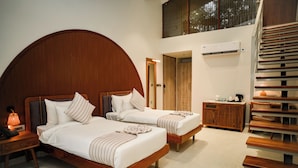 A hotel room with two beds, a wooden headboard, a nightstand with a lamp, a small table with a coffee maker, and a staircase.