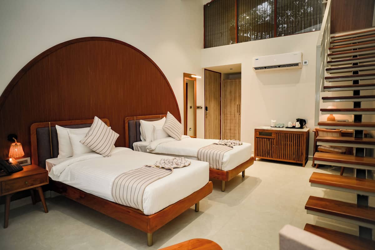 Habitación de hotel con dos camas, cabecero de madera, mesita de noche con lámpara, pequeña mesa con cafetera y una escalera.