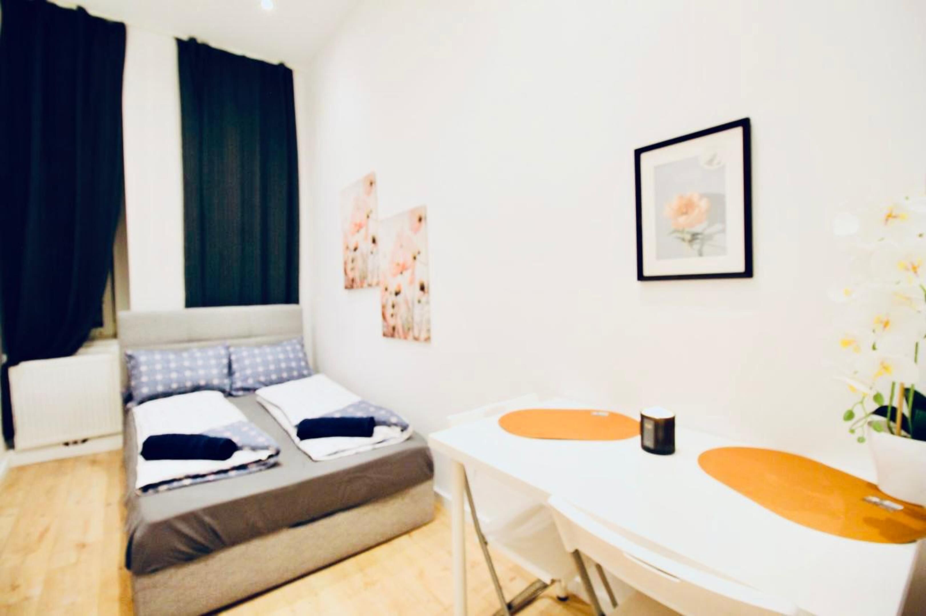 A modern bedroom with a bed, a desk, and a chair. There are curtains, a framed picture, and a vase with flowers.
