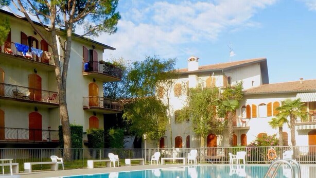 Una piscina con sillas de descanso blancas, rodeada por un edificio con balcones y árboles.