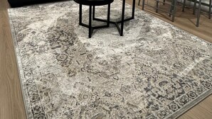 Ein Wohnzimmer mit einer schwarzen Couch, einem runden Couchtisch mit Blumenvase, einem gemusterten Teppich und einem Metallstuhl.