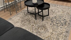 Ein Wohnzimmer mit einer Couch, einem Esstisch mit Stühlen, einem Couchtisch mit Blumenvase, einer Stehlampe, einem Fernseher auf einem Holzschrank und einem gemusterten Teppich.