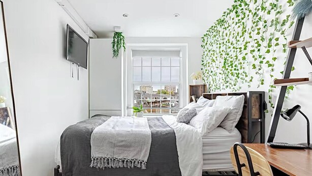 Une chambre à coucher comprenant un lit, un bureau, une chaise, un téléviseur et une fenêtre donnant sur des immeubles.