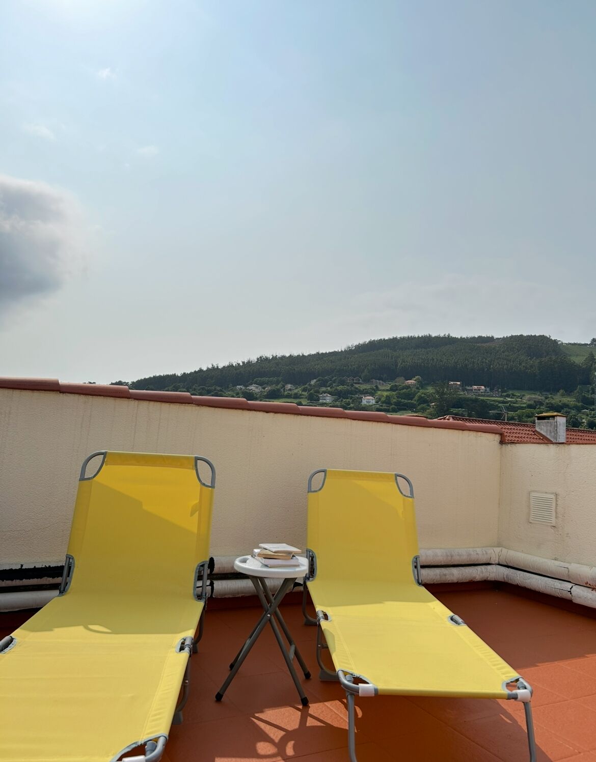 A rooftop terrace with yellow lounge chairs and a small table, overlooking a hilly landscape.