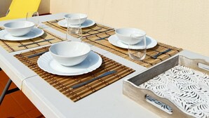 Een eettafel gedekt met witte borden, kommen en bestek op een balkon met een gele stoel, met uitzicht op een bergachtig landschap.
