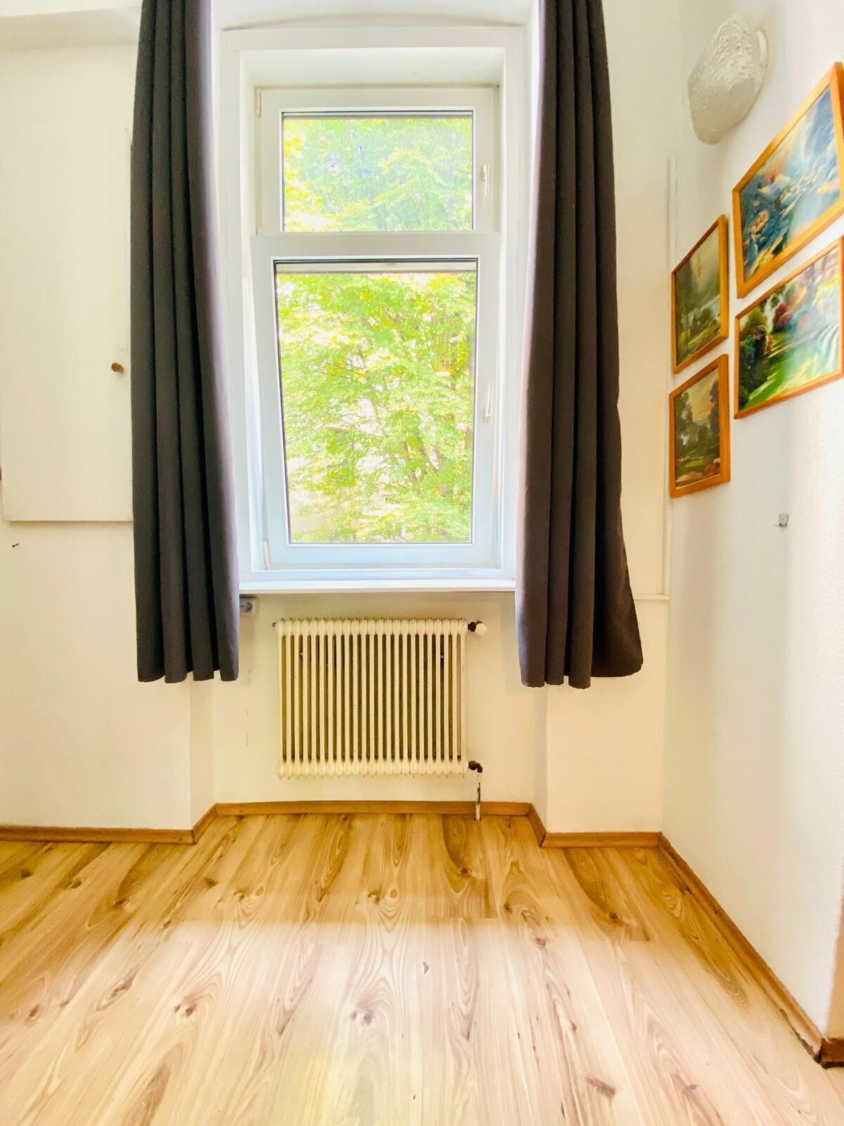 Ein Zimmer mit Fenster, Heizkörper, Holzboden und Wandbild.
