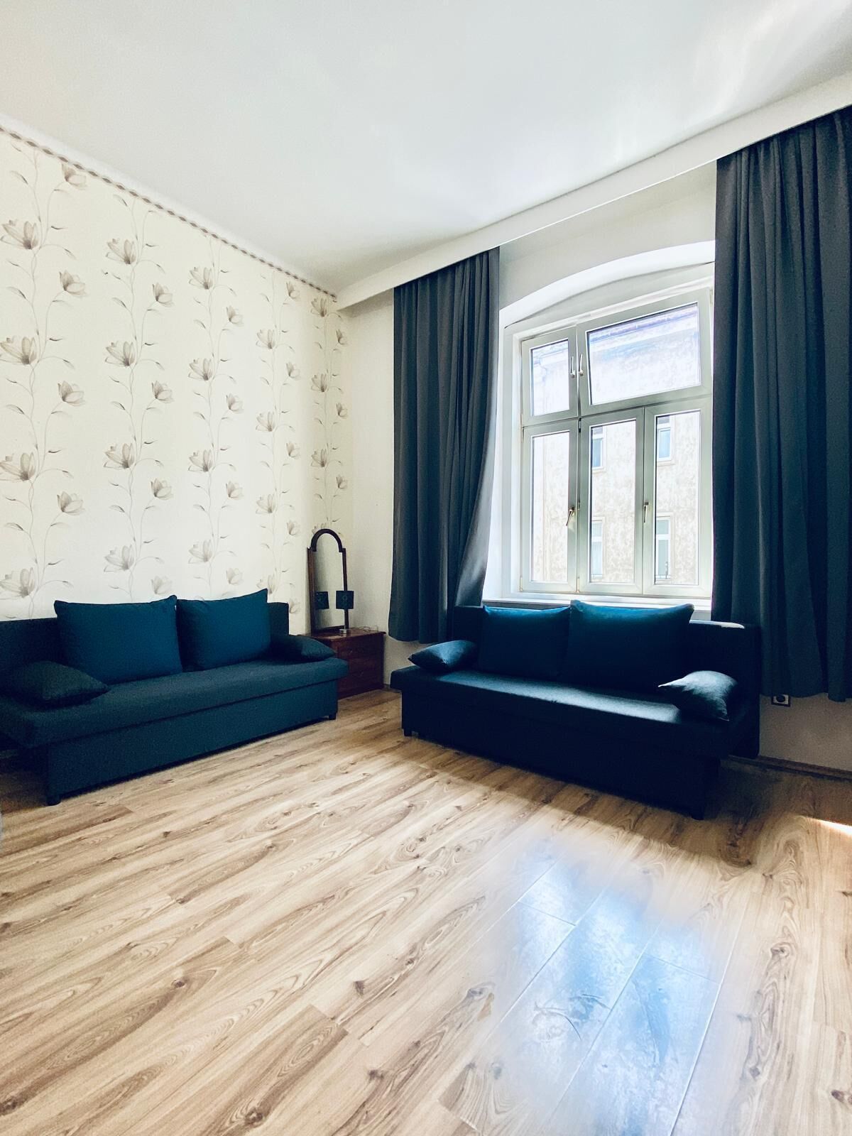 A living room with two dark blue sofas, a wooden floor, a window with curtains, and a wall with a floral pattern.