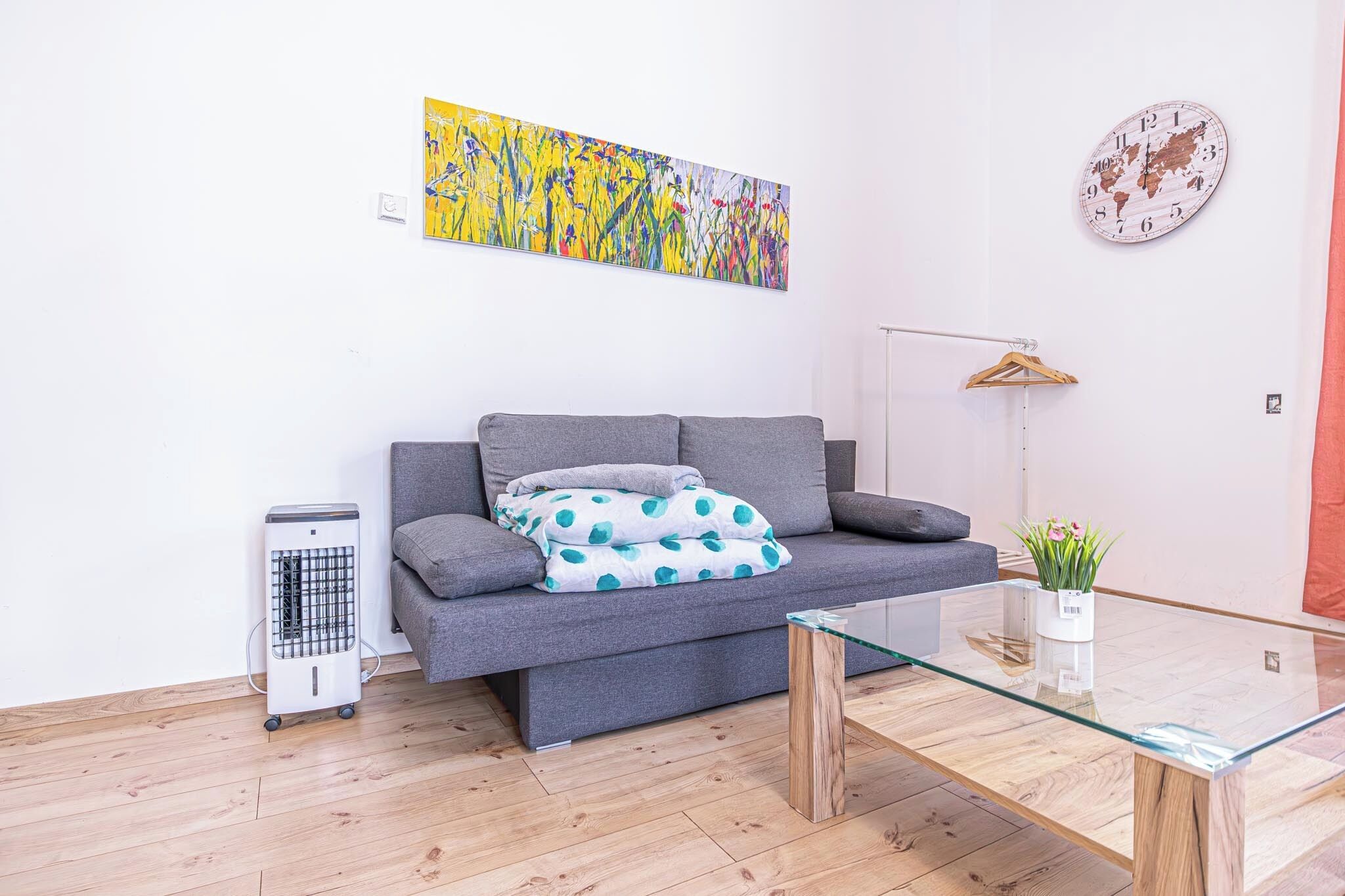 Un salon moderne avec un canapé gris, une table basse en verre, un radiateur, une horloge représentant une carte du monde et un tableau abstrait aux couleurs vives.