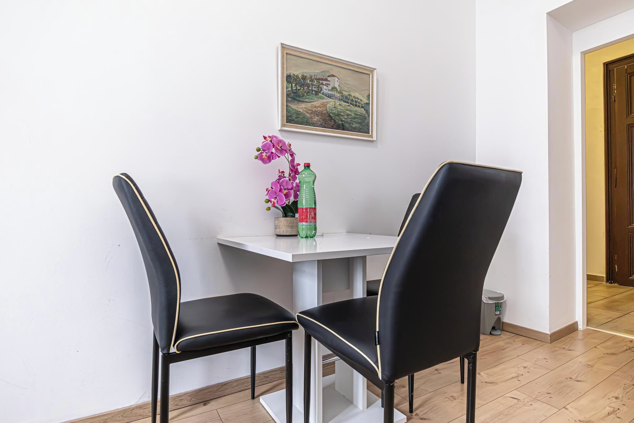 A dining area with a white table and two black chairs with gold trim. A framed picture hangs on the wall above the table.