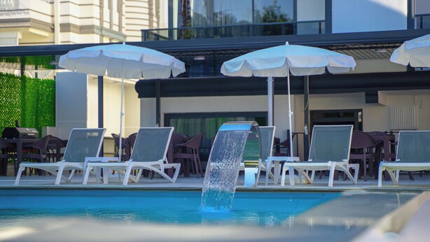Zona de piscina con sillones de descanso y sombrillas, una fuente de agua y un edificio al fondo.
