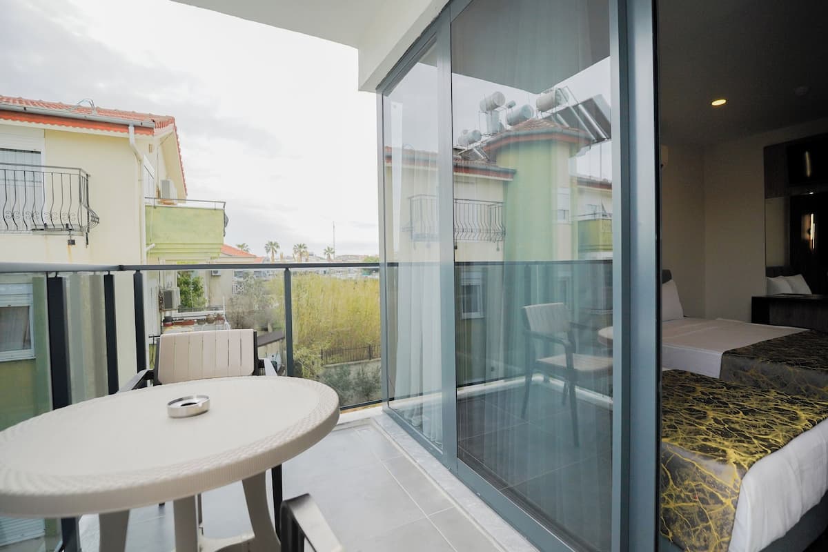 A balcony with a table and chairs, a bed with a patterned bedspread, and a view of neighboring buildings.