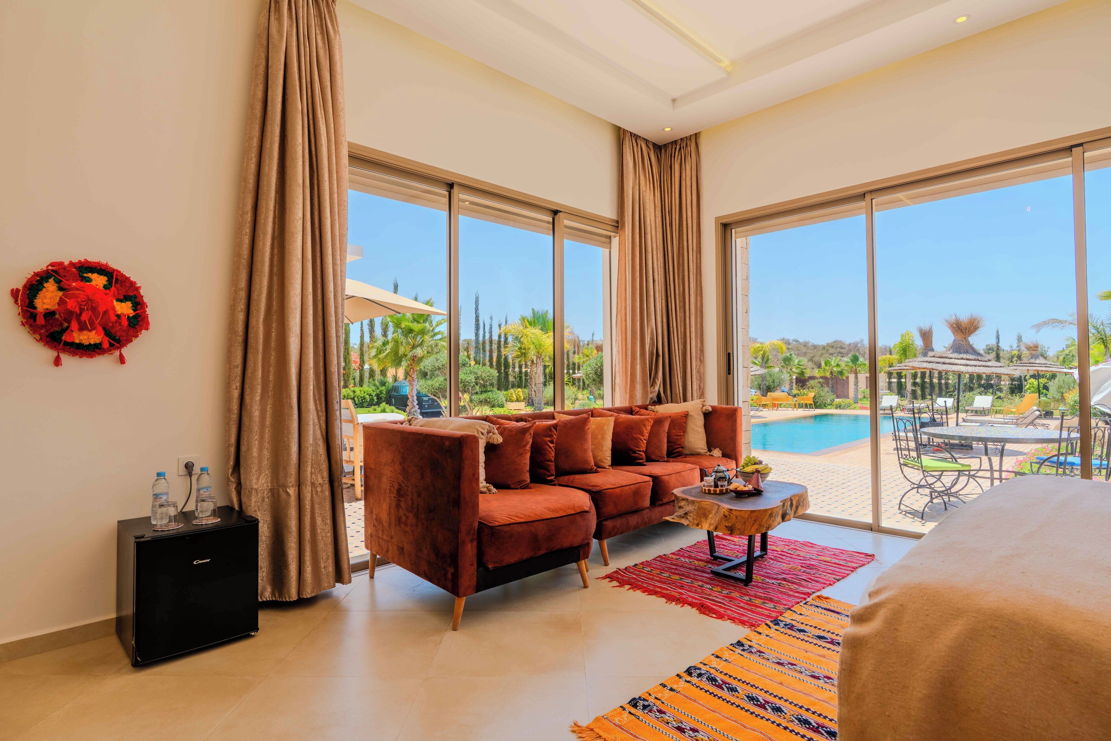 A modern hotel room with a sofa, a small table, and a bed. There is a large window offering a view of a pool and greenery.