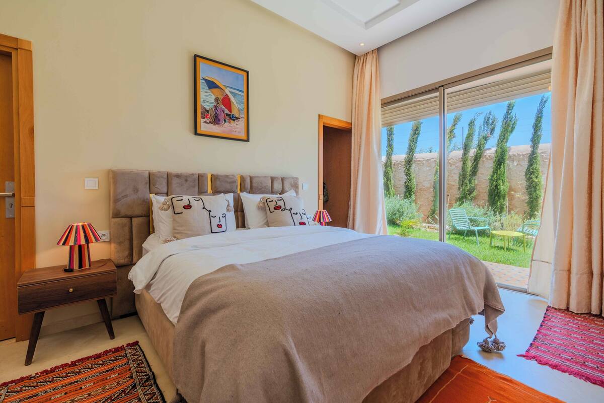 A bedroom with a large bed, a nightstand with a lamp, a painting on the wall, and a view of the garden through a sliding door.