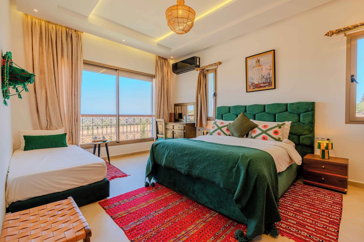 A hotel room with two beds, a balcony, a large window, a green upholstered headboard, and a red patterned rug.
