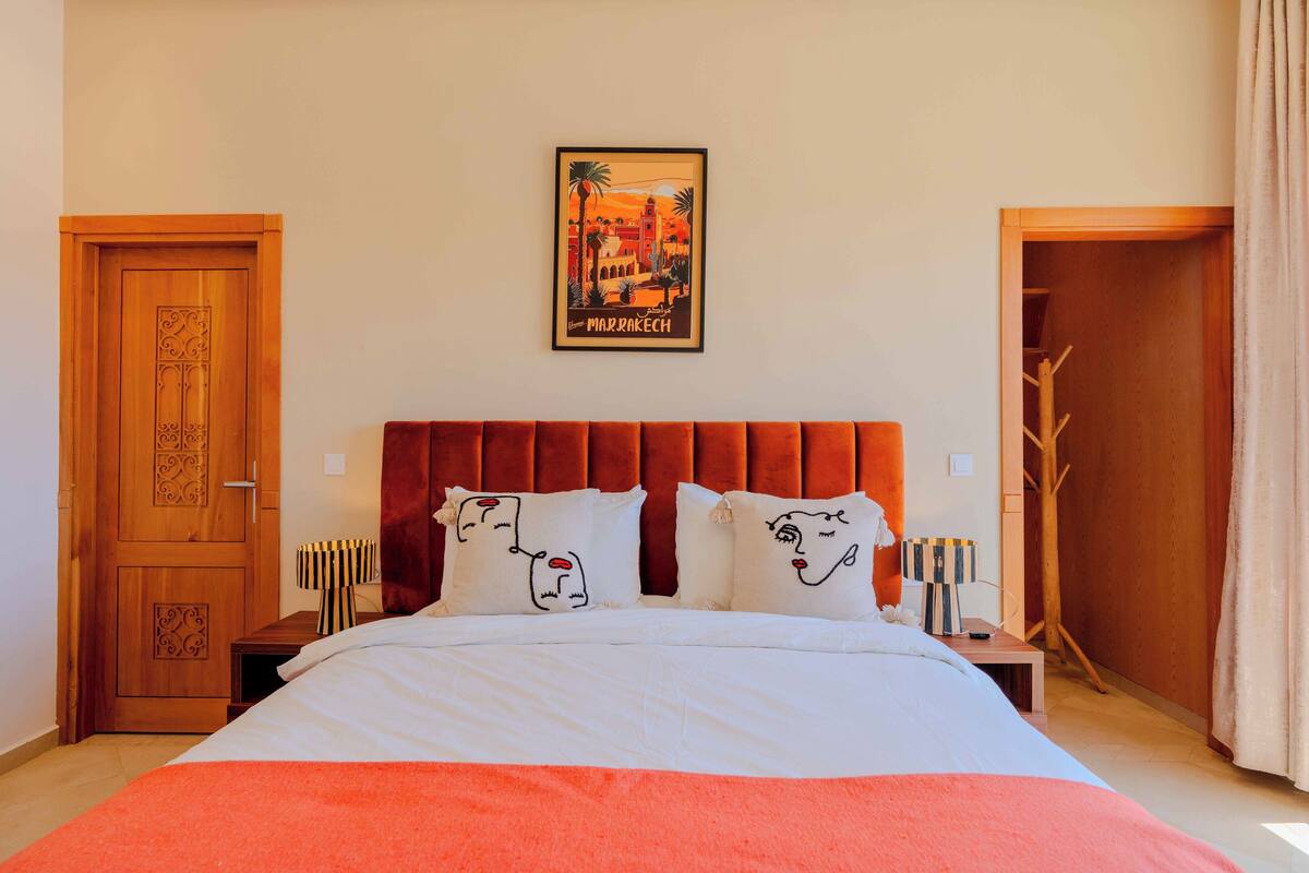 A bedroom with a large bed, two nightstands, a wooden door, and a framed picture of Marrakech on the wall.