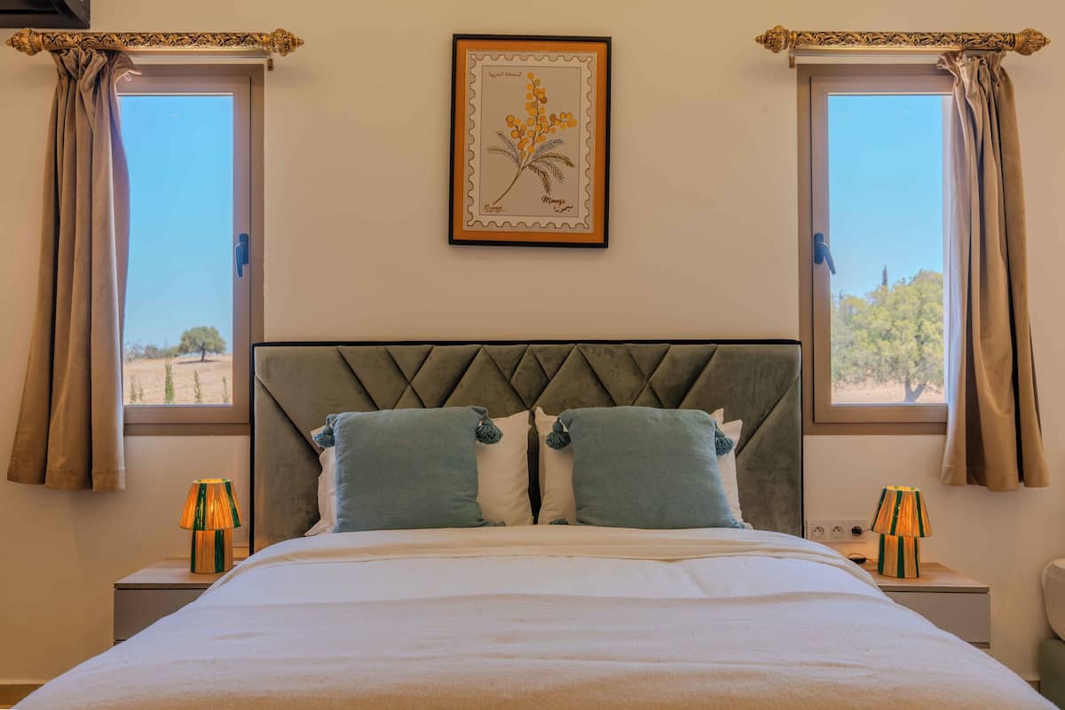 A neatly made bed with two pillows, a framed botanical print on the wall, and two windows with curtains.