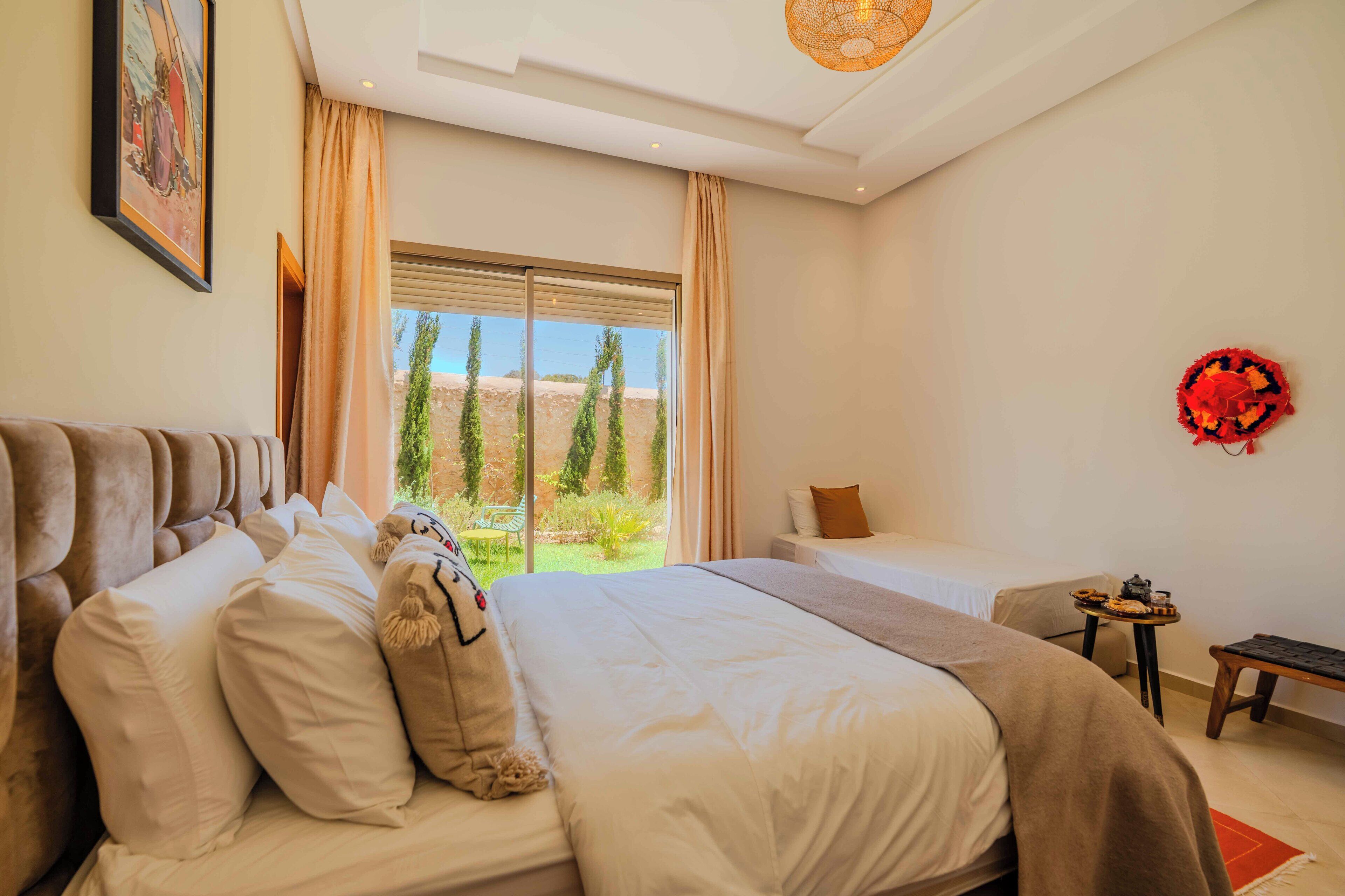 A bedroom with a large bed, a smaller bed, a bench, and a table with a red decorative item.