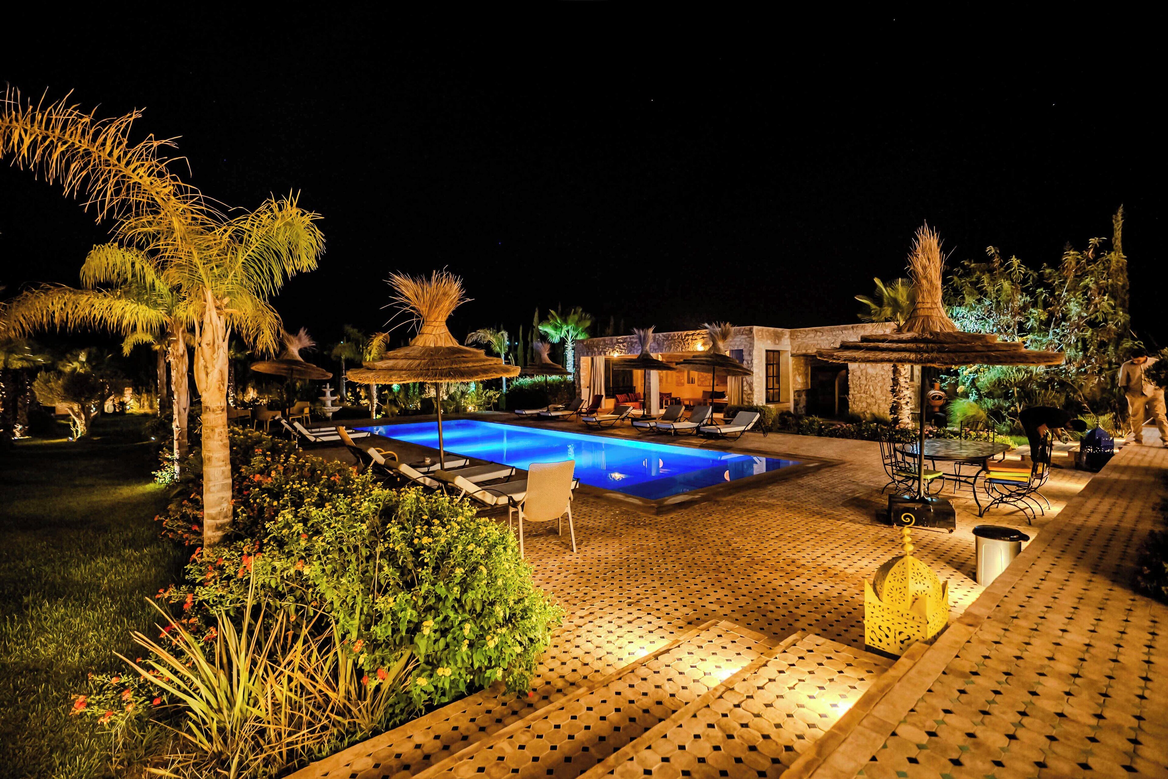 A nighttime outdoor pool area with lounge chairs, a small building, and palm trees.