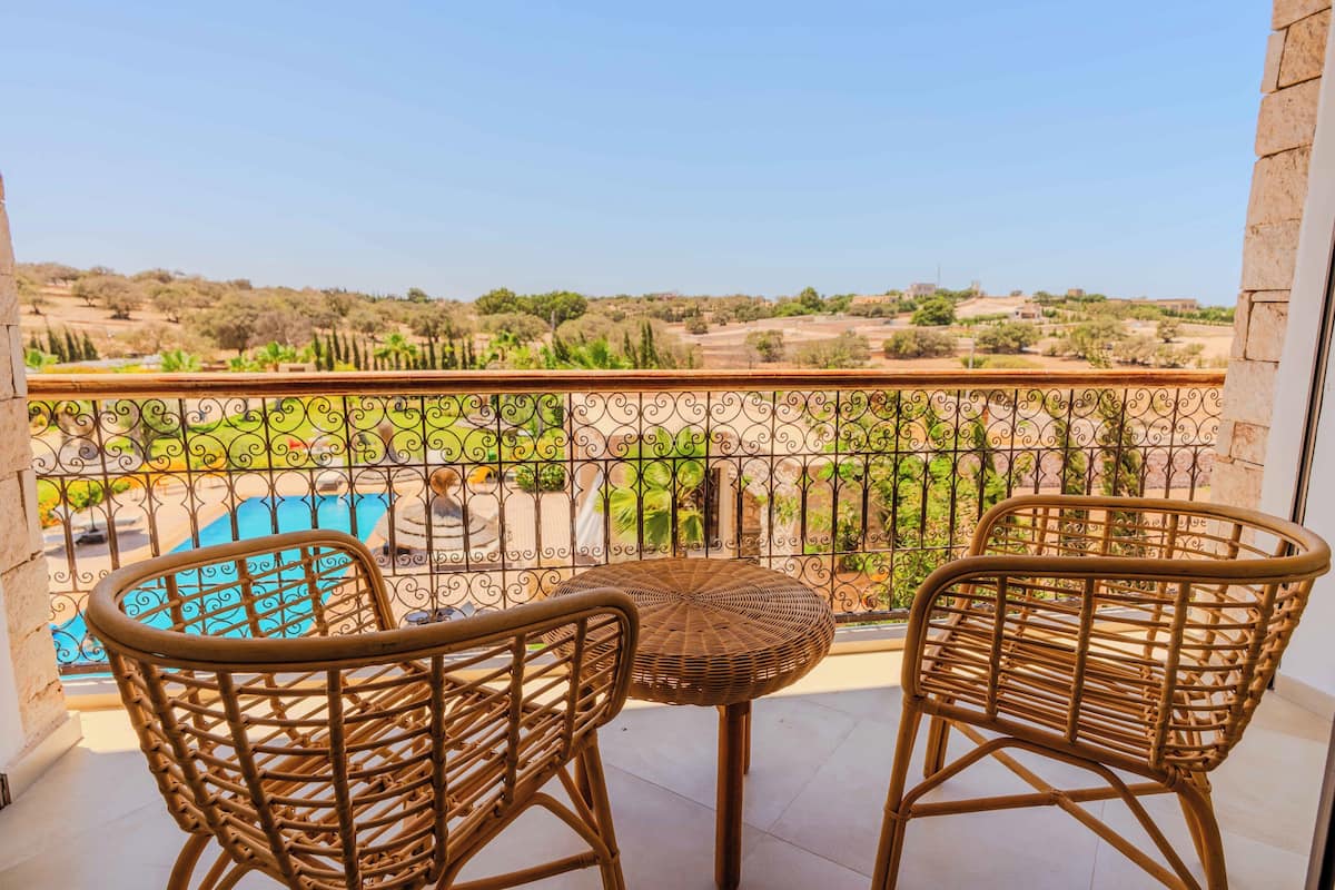A balcony with wicker chairs and a small round table, overlooking a swimming pool and a rural landscape.