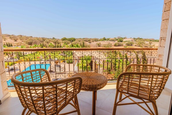 A balcony with wicker chairs and a small round table, overlooking a swimming pool and a rural landscape.