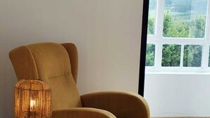 A mustard-colored armchair with a round wooden table beside it, a woven lamp, and a large window with a view of greenery. - Airtegal Casa do Pepe (Cedeira)