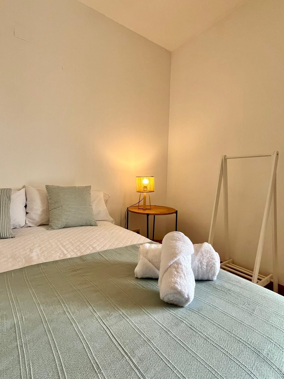 A hotel room with a bed, pillows, a bedside table with a lamp, and a towel rack.