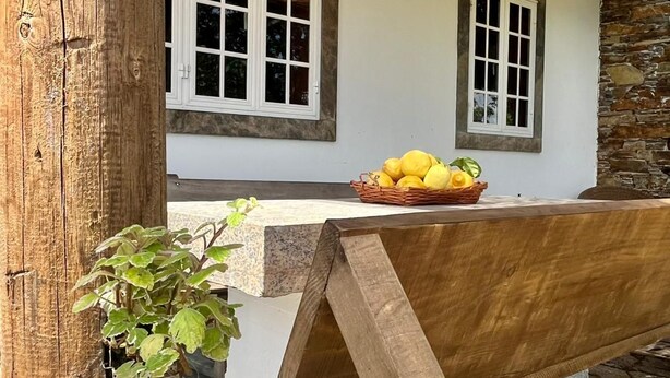 A rustic outdoor seating area with a wooden bench, a woven plant pot, and a basket of yellow fruits on a stone-tiled floor. - Airtegal Casa do Pepe (Cedeira)