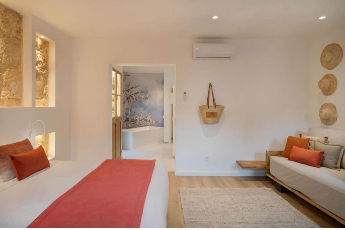 A modern bedroom with a large bed, a sofa, and a small table. There is a wall-mounted air conditioner and a decorative wall hanging.