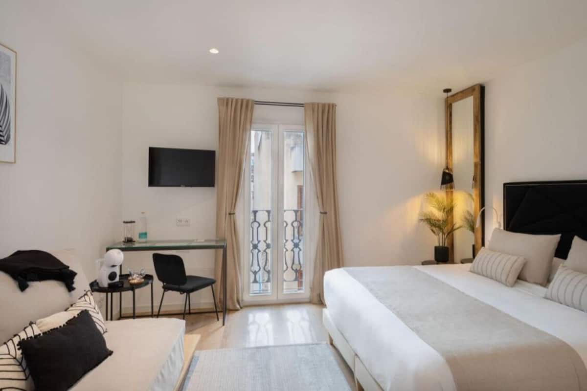 A hotel room with a bed, a desk with a chair, a TV, and a balcony with curtains.
