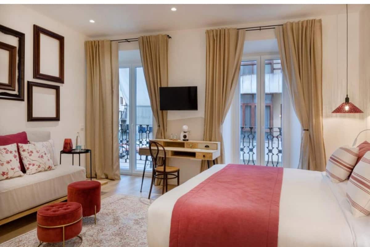 A hotel room with a bed, a desk, a chair, a TV, and a balcony with curtains.