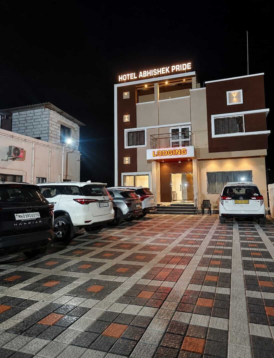 A hotel named 'Hotel Abhishek Pride' with a 'Lodging' sign, multiple parked cars, and a checkered pavement in front.