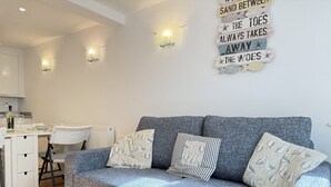 A modern living room with a grey sofa, patterned cushions, a dining area with a white table and chairs, and a wall with a decorative sign. - Penny’s Place – Charming Downalong Apartment Near Beach and Harbour, Perfect for Families (St Ives)