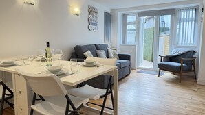 A dining area with a table set for four, a grey sofa, and a wooden chair. - Penny’s Place – Charming Downalong Apartment Near Beach and Harbour, Perfect for Families (St Ives)