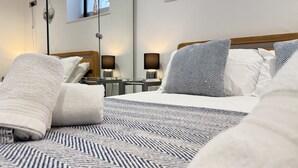 A neatly made bed with a patterned bedspread, white pillows, and a rolled-up towel. There is a bedside table with a lamp and a mirror. - Penny’s Place – Charming Downalong Apartment Near Beach and Harbour, Perfect for Families (St Ives)