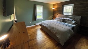 A bedroom with a bed, two windows, a TV mounted on the wall, and a wooden floor.