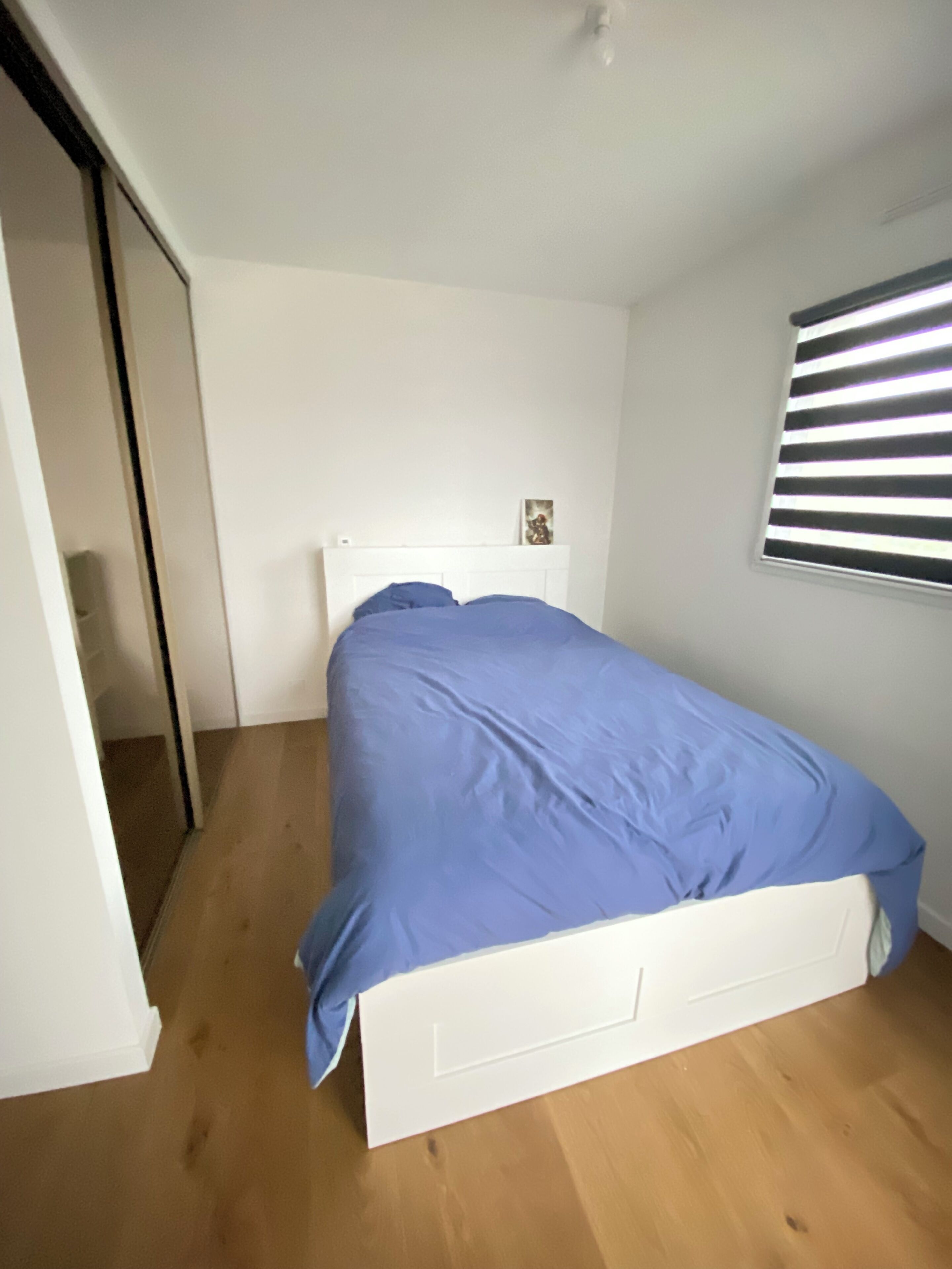 A bedroom with a white bed, a blue bedspread, and a window with horizontal blinds.