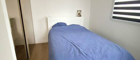A bedroom with a white bed, a blue bedspread, and a window with horizontal blinds.