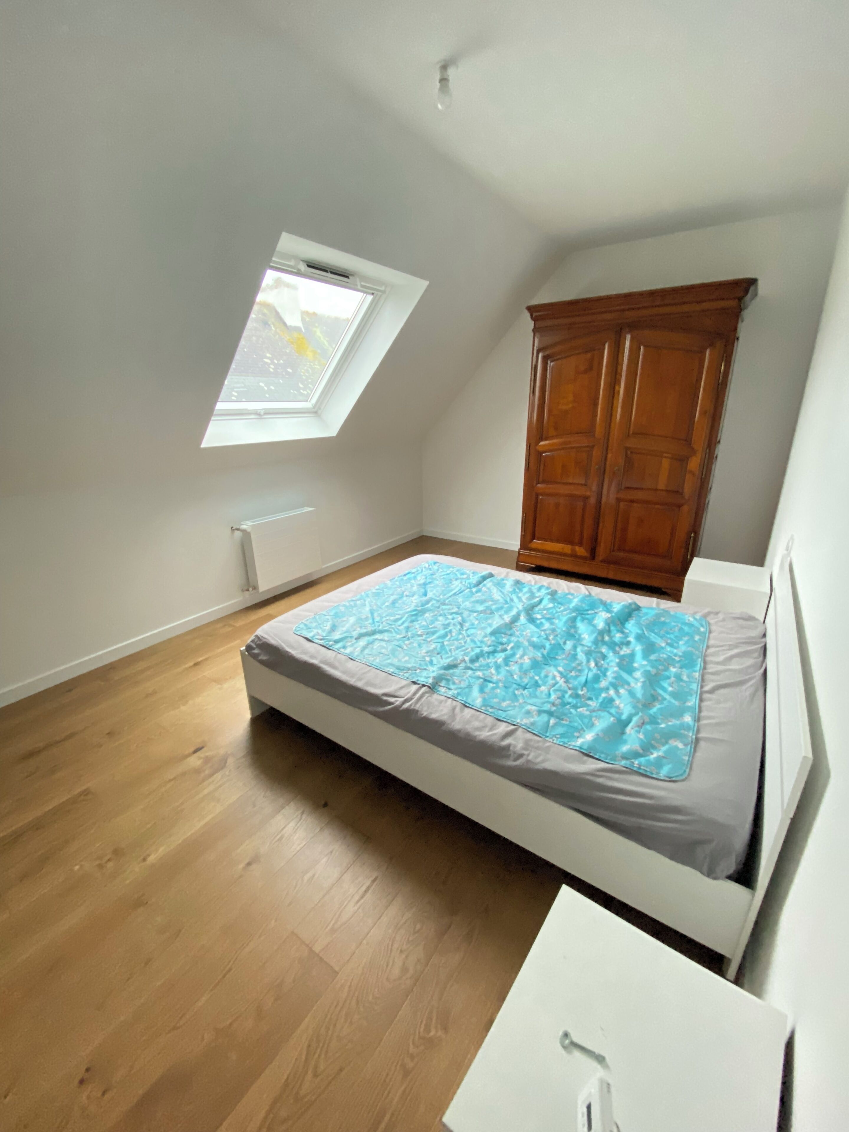 A bedroom with a sloped ceiling, a skylight, a wooden wardrobe, and a bed with a blue blanket.