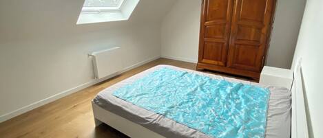 A bedroom with a sloped ceiling, a skylight, a wooden wardrobe, and a bed with a blue blanket.