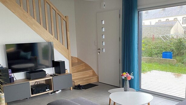 A modern living room with a grey sofa, a flat-screen TV, a wooden staircase, and a view of the outside through a glass door. - A comfortable home in Kérity, just a stone's throw from the sea (Penmarch)