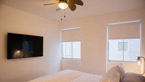 Ein Schlafzimmer mit einem Bett, Deckenventilator, einem Wand-Fernseher und zwei Fenstern mit Jalousien.