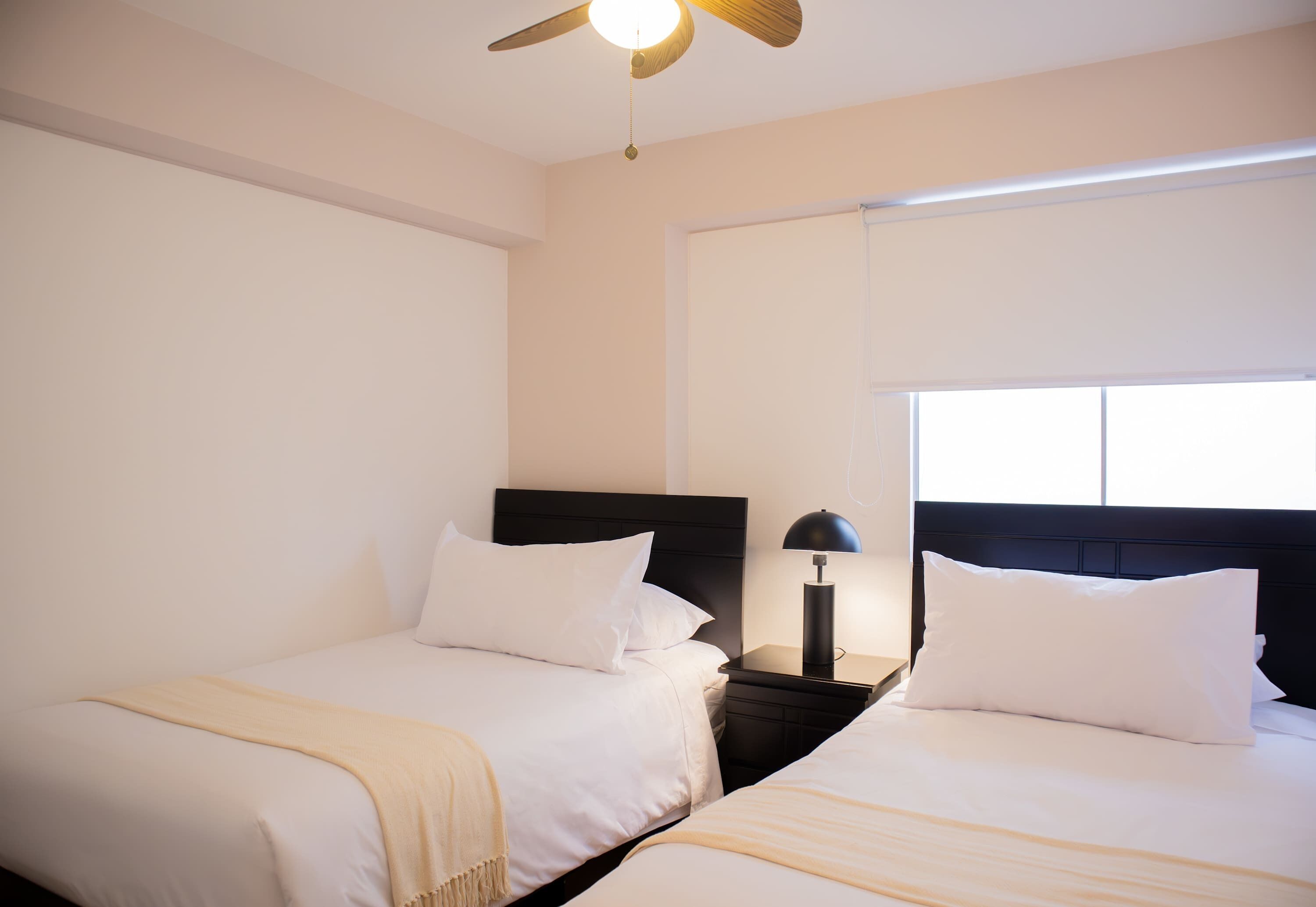 A hotel room with two single beds, a ceiling fan, a nightstand with a lamp, and a window with blinds.