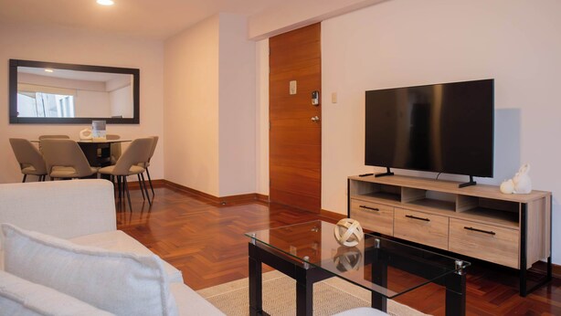 Ein modernes Wohnzimmer mit Flachbildfernseher, Glastisch, Sofa und Essbereich mit Stühlen sowie einer Holztür.